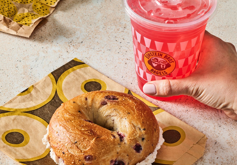 a bagel from einsteins with a woman holding a pink fruit drink